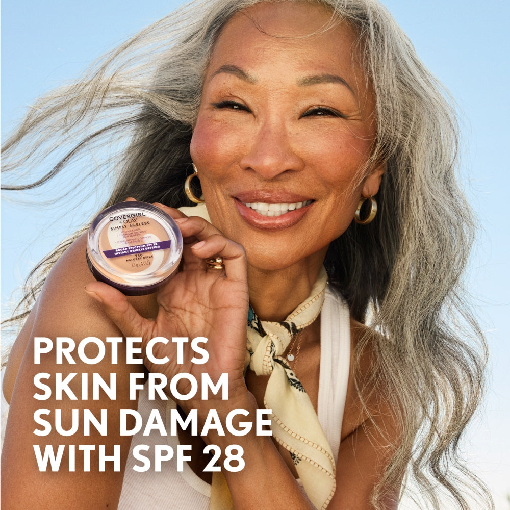 Middle-aged model with long gray hair and gold jewelry holding a container of CoverGirl + Olay Simply Ageless foundation with SPF 28.